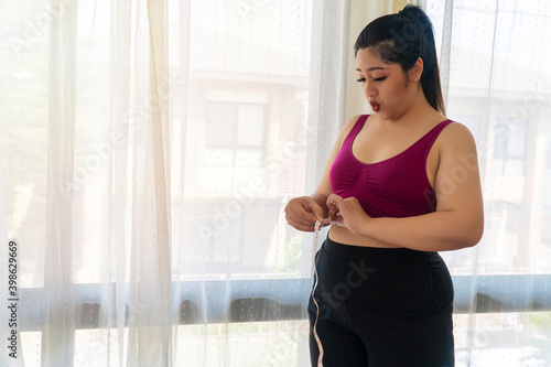 Free Download Obesity unhealthy concept. Overweight woman wear sport bar with measure tape on body pinches fat on her belly large size while standing beside windows feeling unhappy. - GFXLoad