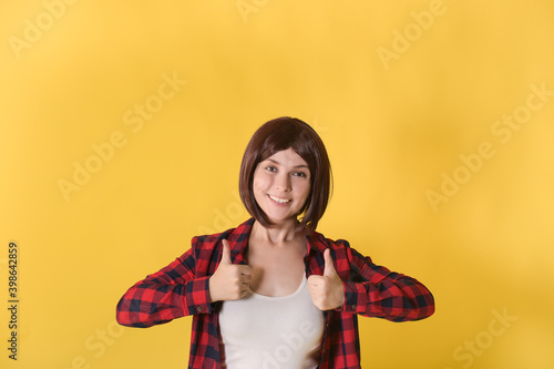 Portrait beautiful young woman standing in checked shirt and gave thumbs up: space for text, well signed sign and looking at the camera on yellow background 
