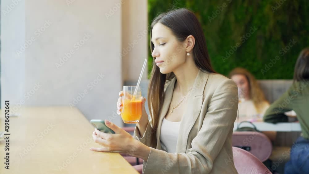 Concentrated brunette with long loose hair in office style wear using smartphone while sitting at city cafe bar with healthy smoothies beverage, at blurred background Concentrated brunette with long loose hair in office style wear using smartphone while sitting at city cafe bar with healthy smoothies beverage, at blurred background