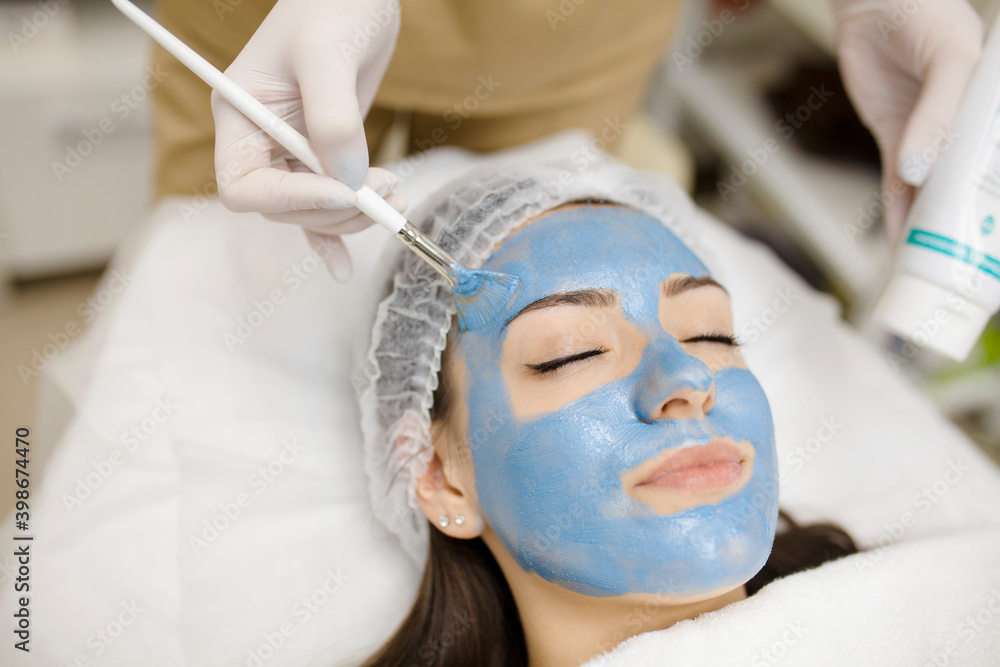 The female cosmetologist applying blue cosmetology mask to young woman face, close-up. Facial cosmetic treatment in beauty clinic.