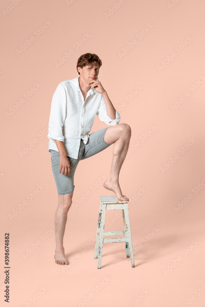 Fototapeta premium Portrait of young man full length wearing white shirt and grey shorts leaned one leg on the chair and looking at camera. Isolated on peach background. 