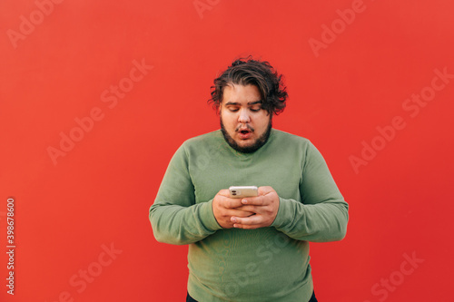 Surprised corpulent young man with a stylish haircut is using a smartphone for texting and chatting, looking concentrated, standing in front of a red wall.