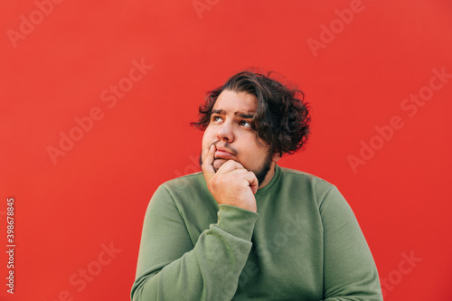 Vászonkép Closeup portrait of a corpulent thoughtful hispanic guy with curly hair, pondering over something, touching his lip showing a thinking gesture, dreaming, looking up