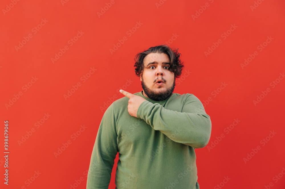 Surprised corpulent hispanic guy wearing a green sweatshirt is pointing his finger backwards on a red background, looking shocked and astonished. Surprised corpulent hispanic guy wearing a green sweatshirt is pointing his finger backwards on a red background, looking shocked and astonished.