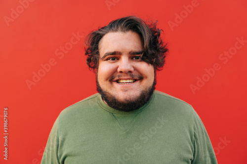 Closeup portrait of a smiling attractive and funny young bearded hispanic man, standing in front of a red wall, looking at the camera with excitement. © bodnarphoto