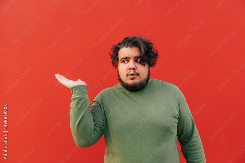 Funny and amusing young hispanic man is displaying the copy space, a red background, with his hand, advertising a product, trying to interest the viewer. Funny and amusing young hispanic man is displaying the copy space, a red background, with his hand, advertising a product, trying to interest the viewer.