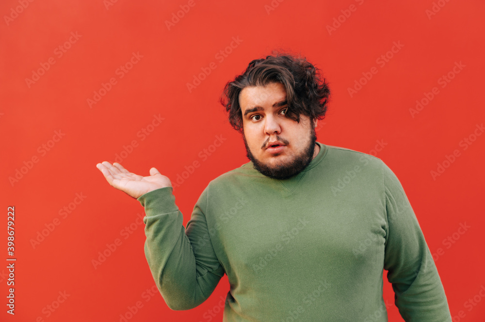 Corpulent bearded hispanic young man is showing on a copy space with his hand, looking at the camera with an interested expression. Corpulent bearded hispanic young man is showing on a copy space with his hand, looking at the camera with an interested expression.