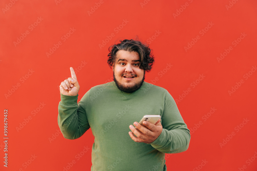Young attractive overweight hispanic man is having an idea, and expressing it with a gesture, pointing his finger up, while using a smartphone. Young attractive overweight hispanic man is having an idea, and expressing it with a gesture, pointing his finger up, while using a smartphone.
