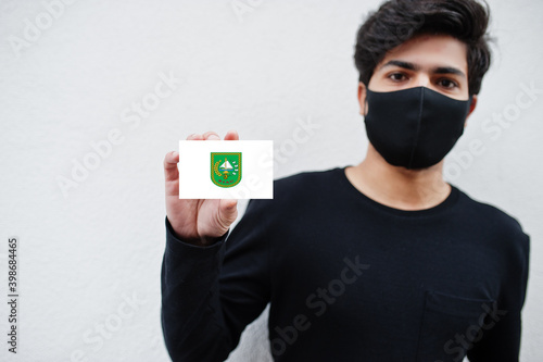 Indonesian man wear all black with face mask hold Riau flag in hand isolated on white background. Provinces of Indonesia coronavirus concept. photo