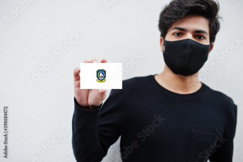 Indonesian man wear all black with face mask hold Riau Islands flag in hand isolated on white background. Provinces of Indonesia coronavirus concept. photo