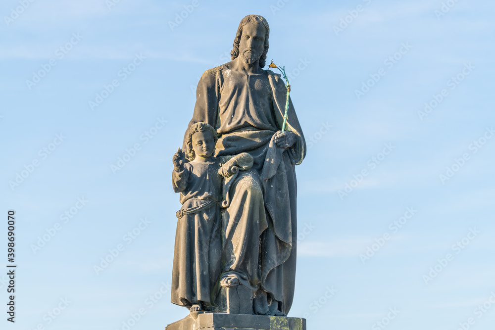 An outdoor statue of St Joseph with a young Christ, on the Charles Bridge, Prague. He holds a flowering rod. The sky behind is blue