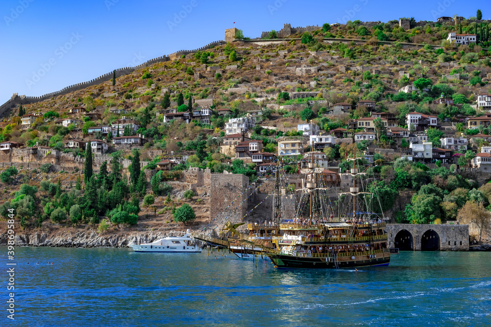 Alanya, Turkey - October 22, 2020: Pirate ship for parties in Mediterranean Sea against the background of Alanya old town. Seascape with ruins of ancient buildings, medieval wall and modern cottages