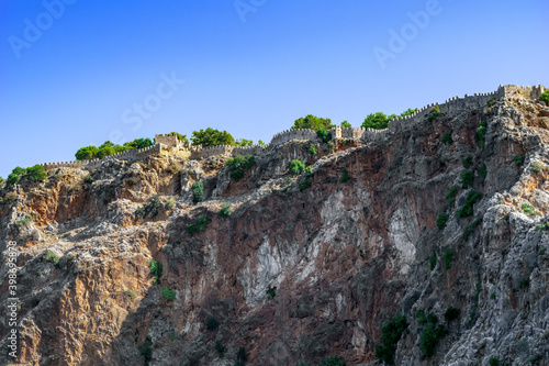 Ancient stone wall of Alanya castle (Turkey) at the top of a steep cliff - bottom view. Beautiful natural landscape with brown rocks, clear blue sky, green plants and an old fortress