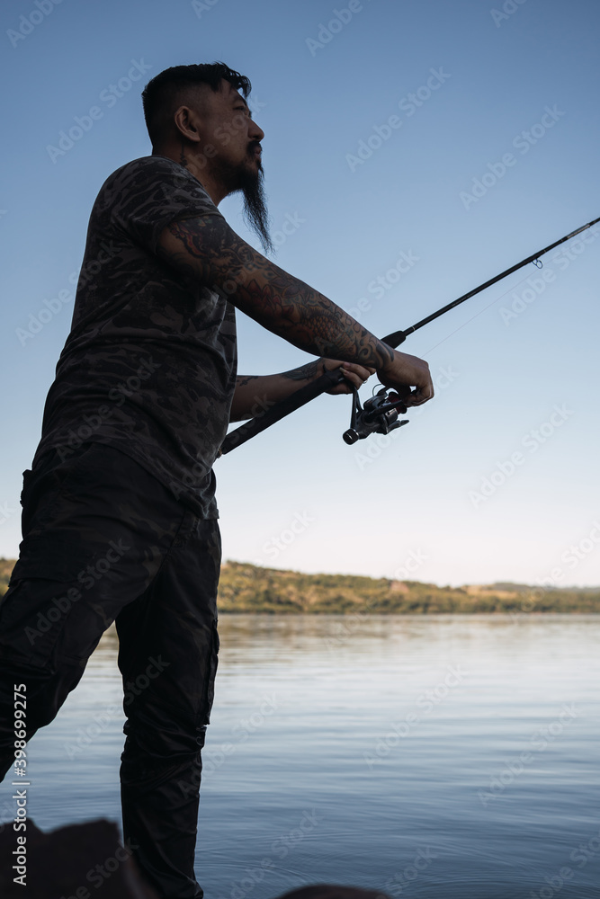 Asian man with beard and shirt camouflaged in the river - Young fisherman casting his fishing rod during a sunset fishing session in front of the river. Asian man with beard and shirt camouflaged in the river - Young fisherman casting his fishing rod during a sunset fishing session in front of the river.