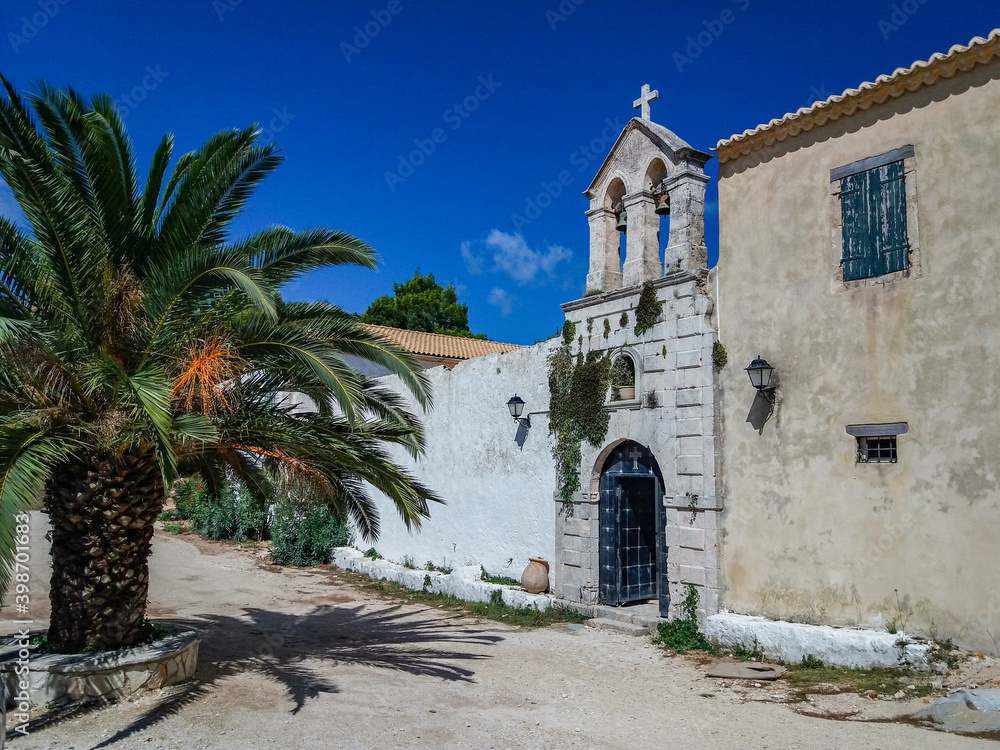 Old mediterranean church made of stone with a cross, on the top at the village sqare with a palm tree and blue sky in background in Greece
