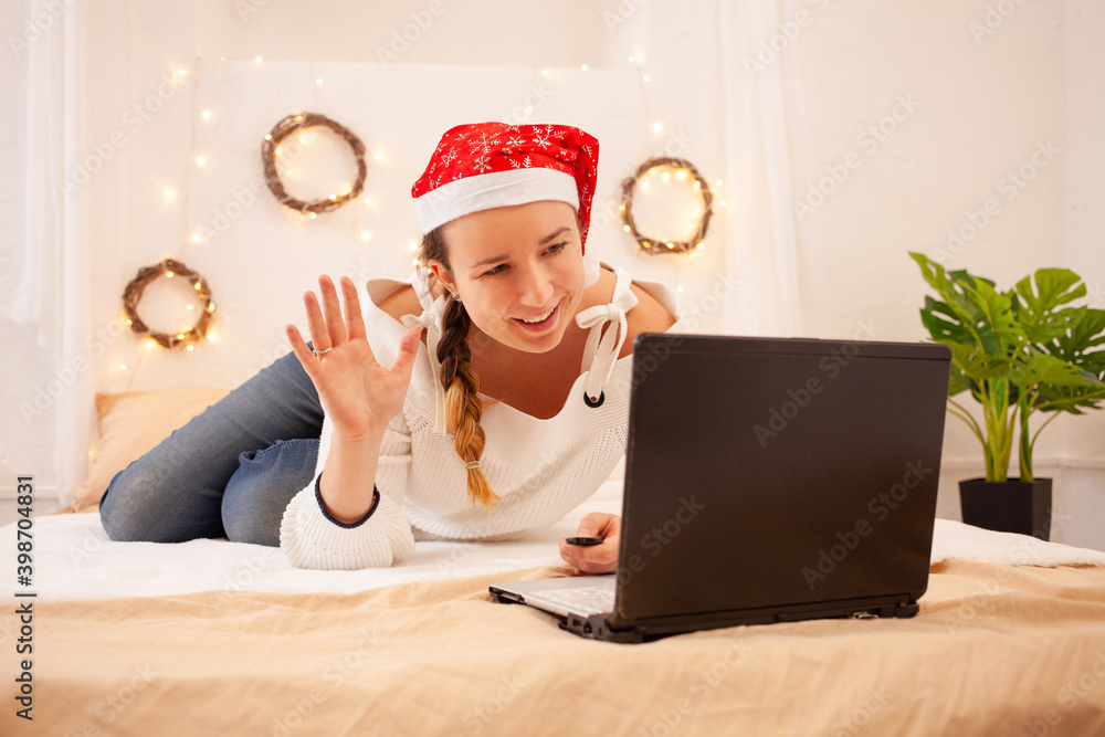 A beautiful Woman smiles and waves her hand to her laptop. A girl in a hat at Christmas congratulates her relatives by video link. New Year in quarantine, a safe holiday from coronavirus. A beautiful Woman smiles and waves her hand to her laptop. A girl in a hat at Christmas congratulates her relatives by video link. New Year in quarantine, a safe holiday from coronavirus.