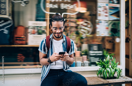 Smiling dark skinned male millennial checking income messages on smartphone standing on urban blurred background, positive african american guy influencer blogger share media content with followers