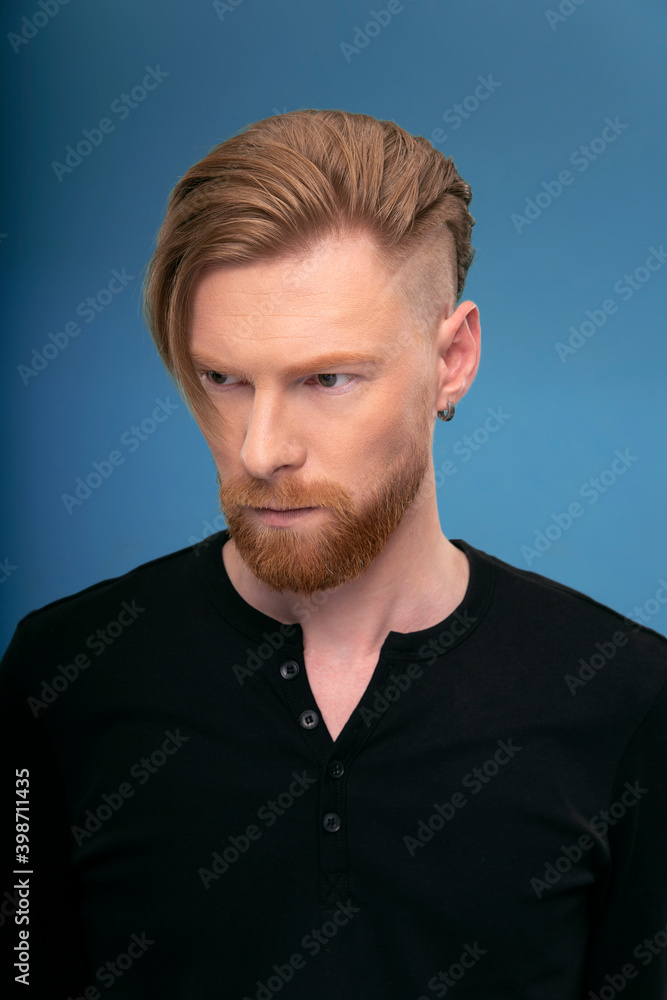 Fototapeta premium Handsome redhaired bearded man, wearing black shirt, posing frontal in studio, looking seriously, over blue background.