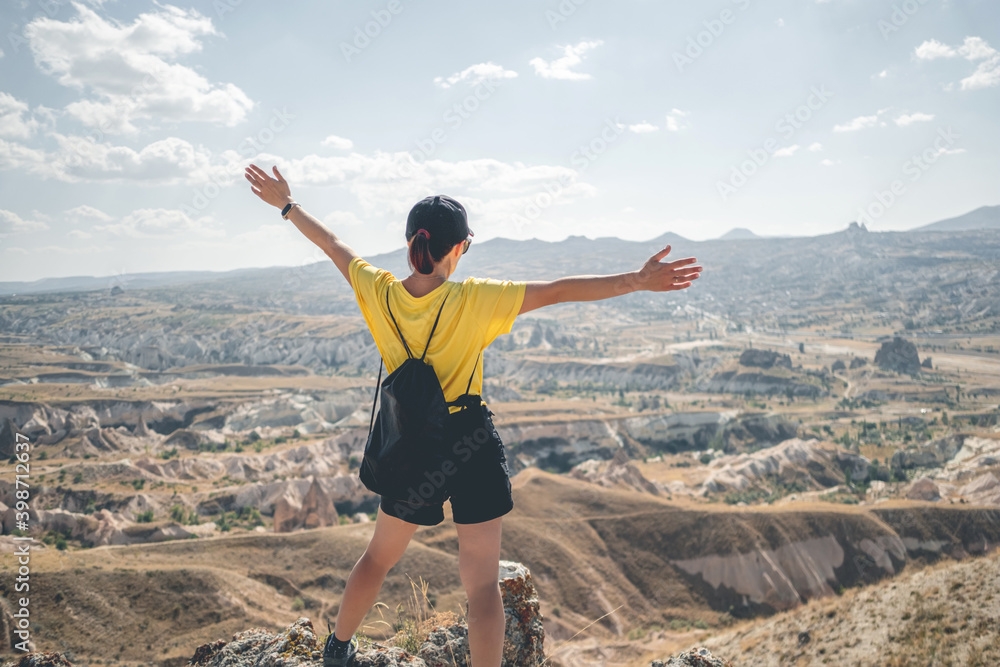 Woman tourist with backpack standing backwards on mountain top in Cappadocia with open arms, Turkey