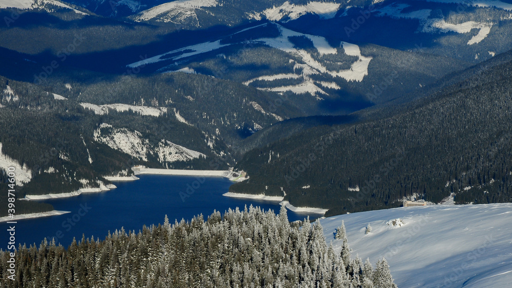 Vidra lake during winter season. The spruce forests are covered with a layer of fresh snow, the higher altitude trees are frozen. View from the ski piste Transalpina Ski resort. Carpathia, Romania
