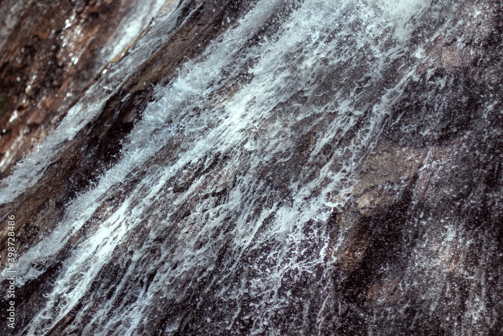 Texture of a waterfall rushing down the mountain steep slope. Falling down stormy water from high rocky cliff for background design. Active fine severe splashes of nature shower on abrasive wall rock