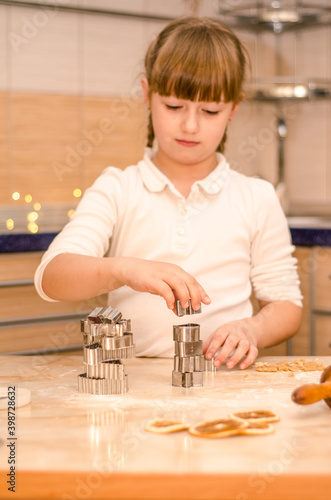 Cute happy girl learns how to cook Christmas gingerbread cookies in bright kitchen, builds turret out of cookie cutters