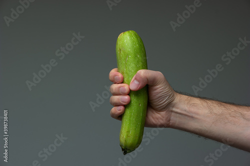 Caucasian male hand holding a phallic piece of fresh green ripe zucchini close up shot isolated on gray