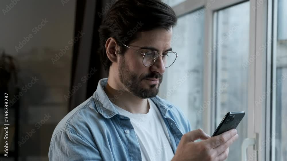 Young happy man standing near window indoors at home while using mobile phone Young happy man standing near window indoors at home while using mobile phone