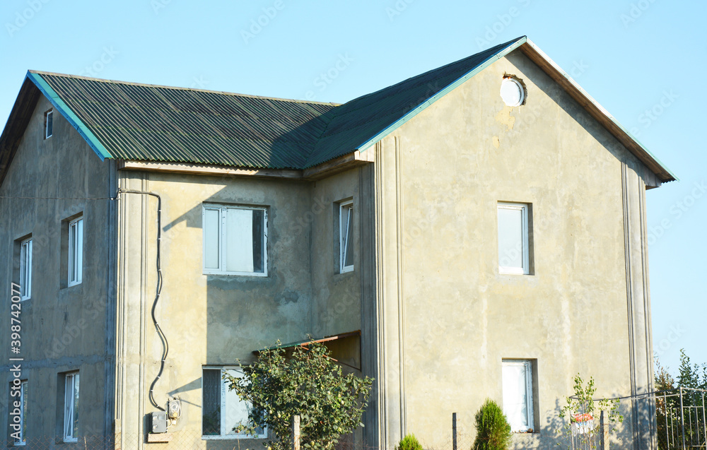 An unfinished stucco facade of a large house that needs rendering, painting and renovation with asphalt shingled roof, rain gutters, small round attic windows and outdoor electricity conduit, cables. An unfinished stucco facade of a large house that needs rendering, painting and renovation with asphalt shingled roof, rain gutters, small round attic windows and outdoor electricity conduit, cables.