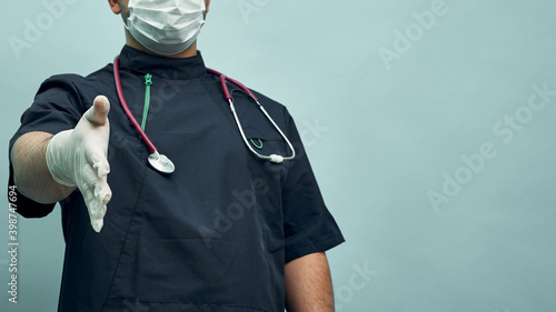 Young doctor man stretches out his hand for a handshake. Protective mask and gloves, professional clothing. Greetings in the face of a pandemic.