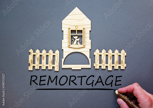 Remortgage symbol. Hand writing 'remortgage' on grey background. Model of a wooden house. Male hand, wooden fence. Copy space. Business and remortgage concept. photo