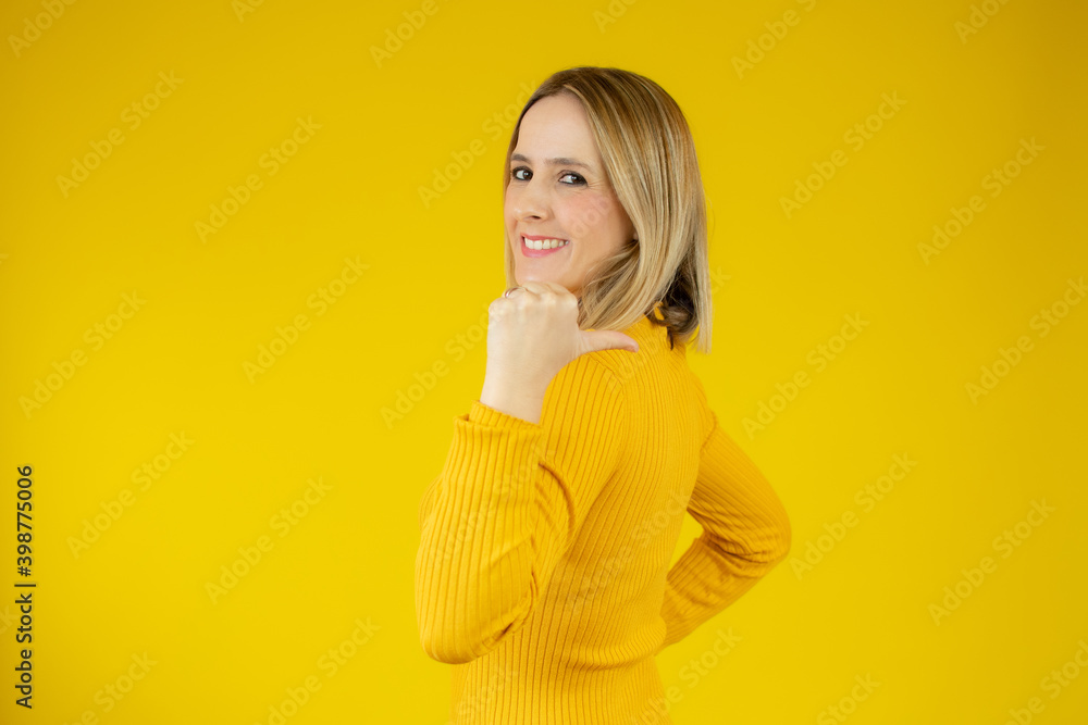 Smiling woman wearing casual sweater pointing thumb side. Isolated portrait on yellow.