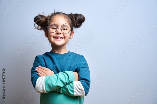 Portrait of cute swarthy caucasian little girl with glasses, arms crossed and funny hairstyle, beautiful happy child smiling and laughing looking at camera isolated in studio