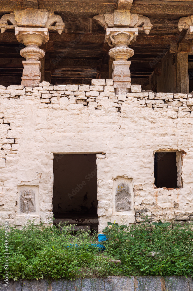 Fototapeta premium Hampi, Karnataka, India - November 4, 2013: Hampi Bazar street. Historic ruinous empty bulding with decorated roof holding pillars and frescos on white painted wall. Green foliage in front.