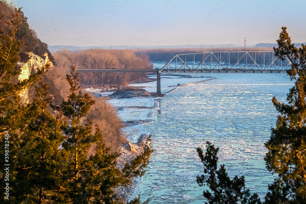 View of Missouri River at sunset with ice floes in the water; river bluffs on the left, highway bridge in the distance; sky in background