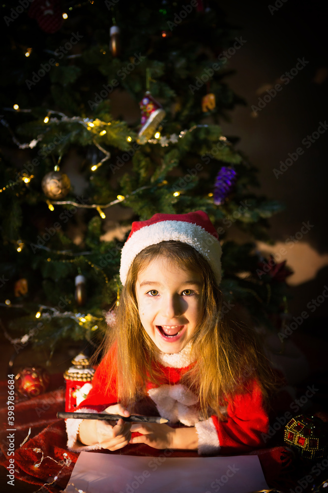 Little girl in a Santa hat and red dress under Christmas tree is dreaming, waiting for the holiday, lying on a plaid blanket. A letter on piece of paper, gifts. New year, Christmas. defocus lights Little girl in a Santa hat and red dress under Christmas tree is dreaming, waiting for the holiday, lying on a plaid blanket. A letter on piece of paper, gifts. New year, Christmas. defocus lights