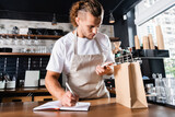handsome, barista in apron writing in notebook while looking at smartphone near paper bag on bar counter