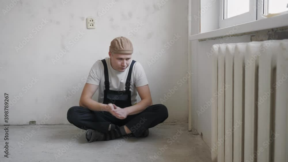 A man in construction overalls is resting sitting on the floor near the wall after the work done to repair the apartment. High quality 4k footage A man in construction overalls is resting sitting on the floor near the wall after the work done to repair the apartment. High quality 4k footage