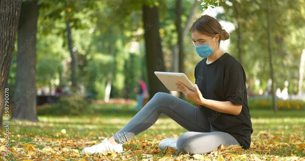 Young girl in medical protective mask sits on the lawn in autumn city park and looks photos in tablet computer. Cinema 4K 60fps video Young girl in medical protective mask sits on the lawn in autumn city park and looks photos in tablet computer. Cinema 4K 60fps video