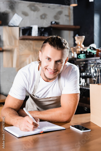 Barista looking at camera while writing in notebook near smartphone with blank screen on bar counter