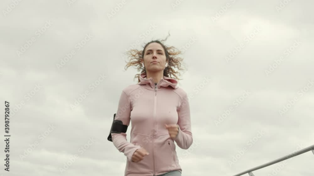 Medium shot of young good-looking Caucasian sporty woman wearing sportswear and wireless headphones having a run along embankment in downtown Medium shot of young good-looking Caucasian sporty woman wearing sportswear and wireless headphones having a run along embankment in downtown