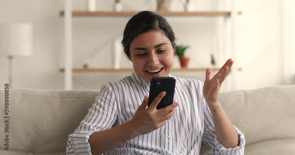 Pretty Indian woman sit on couch with modern smartphone read sms celebrate success. Concept of on-line game fan betting victory, gambling, fantastic unbelievable news receiving moment, candid emotions Pretty Indian woman sit on couch with modern smartphone read sms celebrate success. Concept of on-line game fan betting victory, gambling, fantastic unbelievable news receiving moment, candid emotions