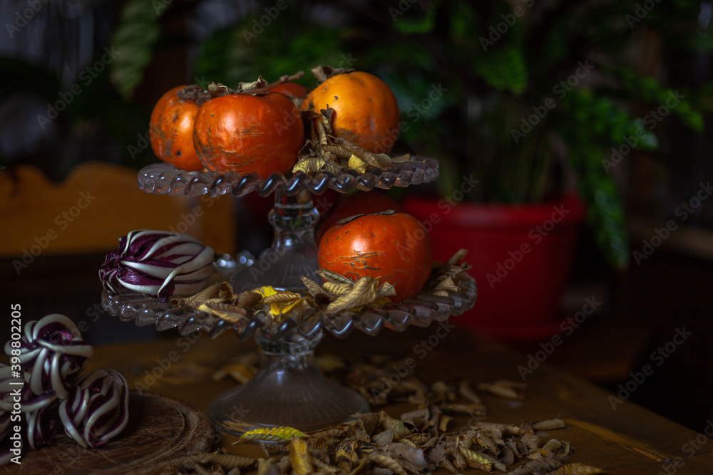 Fruit holder on wooden table with persimmon and red radicchio with a base of dry yellow leaves. Green plants background