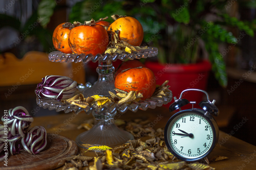 Fruit holder on wooden table with persimmon and red radicchio with a base of dry yellow leaves. Green plants background. Presence of an old alarm clock on the right side