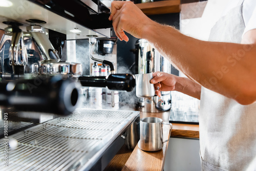 Partial of barista holding metallic milk mug near steamer of coffeemaker on blurred foreground