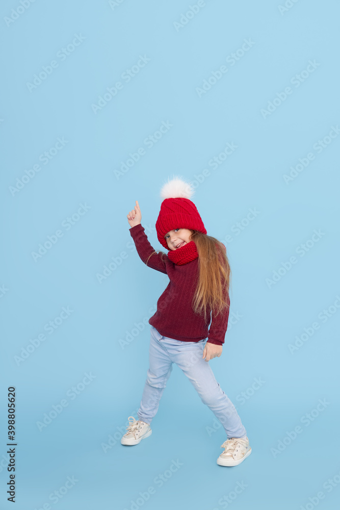 Winter portrait of happy child girl in a knitted red hat, snood and sweater points up. Blue background. childrens winter entertainment Winter portrait of happy child girl in a knitted red hat, snood and sweater points up. Blue background. childrens winter entertainment
