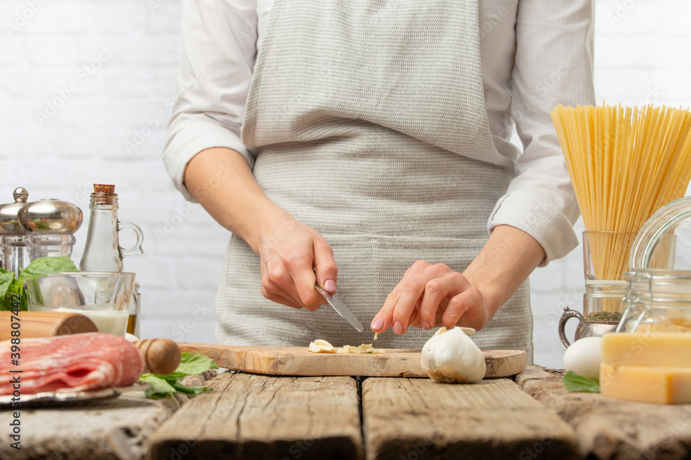 Professional chef cuts with knife garlic on wooden board for cooking pasta alla carbonara. Backstage of preparing traditional italian dish on white background. Frozen motion. Cookbook illustration.