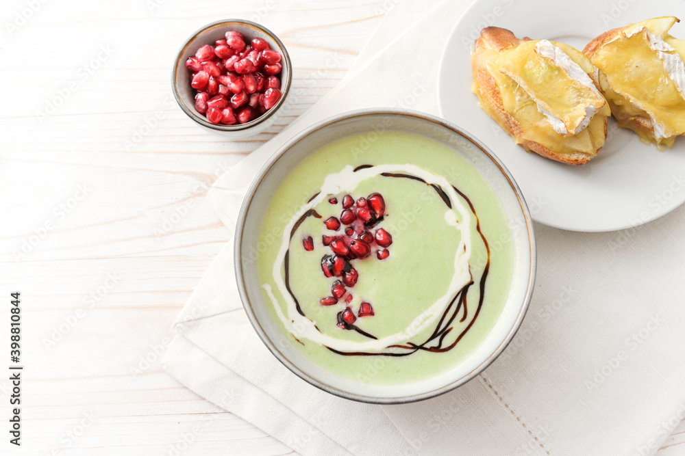 Festive soup of green apple and peas with pomegranate seeds and baked camembert crostini in a bowl on a white wooden table, copy space, high angle view from above