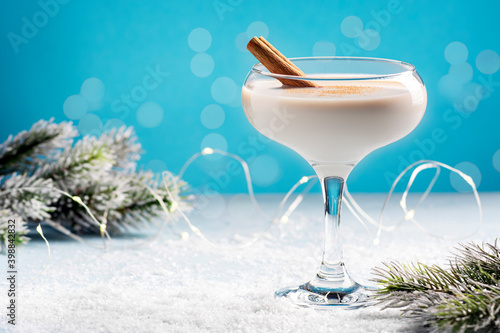 Winter hot eggnog in a beautiful glass on a blue background covered with snow, near a garland and spruce branches, bokeh, horizontal orientation