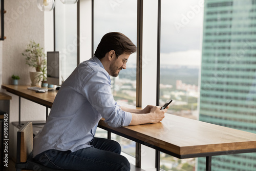 Young Caucasian man sit at desk in modern building use smartphone browsing internet on gadget. Happy millennial male look at cellphone screen, texting messaging online. Technology concept.